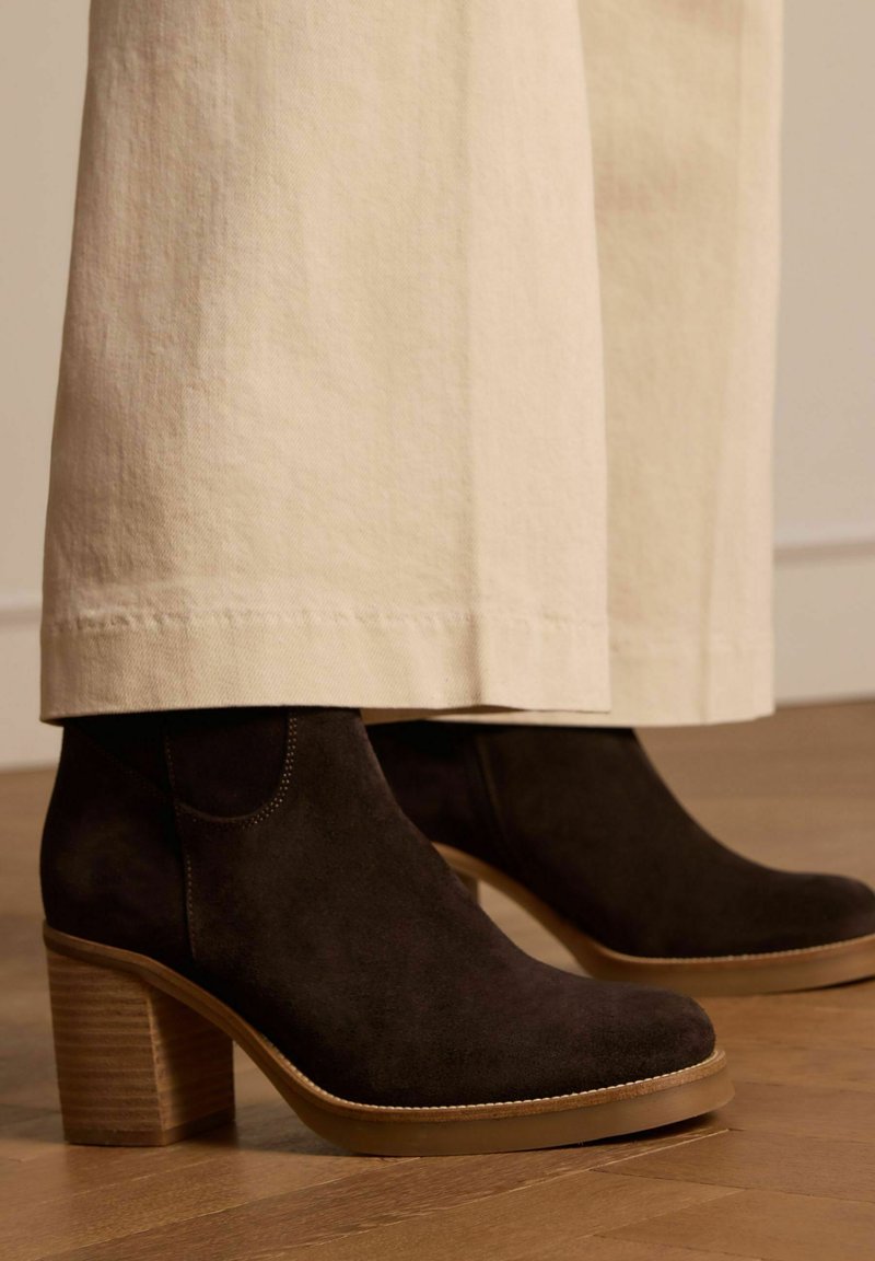 Donkerbruin suède enkellaarsjes met een chunky houten hak en een gladde rubberen zool. Ze hebben subtiele stiksels langs de bovenrand.