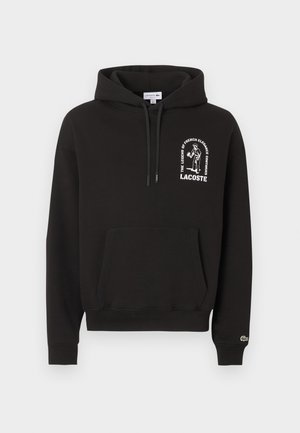 Sweat à capuche noir avec poche avant, motif blanc et texte "La légende de l'élégance française continue" au-dessus du logo Lacoste sur la poitrine.