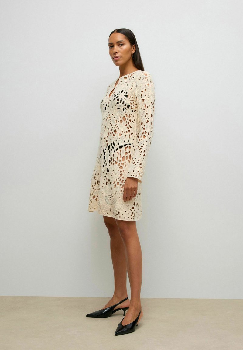 Femme portant une robe en dentelle crochet beige sur des sous-vêtements noirs, assortie à des talons aiguilles noirs à bride arrière, debout contre un mur uni.