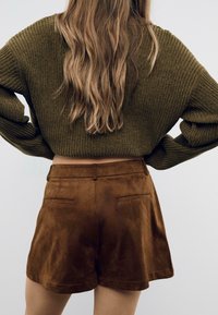 Donna con lunghi capelli mossi, che indossa un maglione a coste verde oliva e pantaloni corti in suede marrone a vita alta, con le mani sui fianchi.