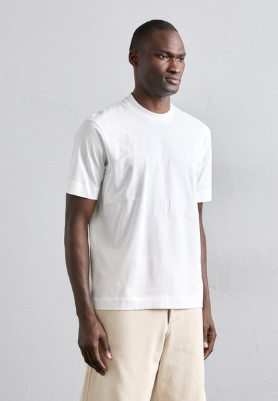 T-shirt en coton blanc à manches courtes et col rond, avec une texture lisse et un design minimaliste, porté avec un pantalon beige.