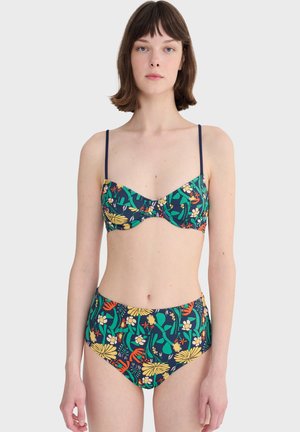 Femme portant un maillot de bain deux-pièces à motifs floraux verts, jaunes, oranges et blancs sur un fond bleu foncé, se tenant devant un fond gris uni.