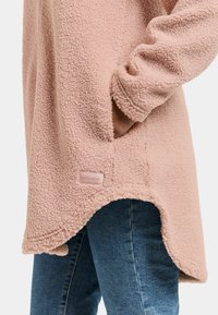 Pull-over en molleton rose avec un tissu texturé, des poches latérales et un ourlet arrondi. Il comporte une petite étiquette de marque au bas.
