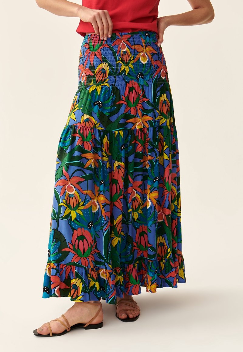 Femme portant une longue jupe fleurie colorée avec une taille smockée et un haut rouge, debout avec une main sur la taille et des sandales pieds nus.