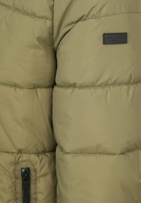 Olijfgroene pufferjack met een gladde textuur, horizontale stiksels en een ritssluiting zak. Heeft een zwart merkembleem op de borst.