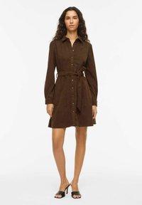 Pruun sametkostüümkleit krae, pikkade varrukate, nööpidega kinnituse, vöökohal sõlmitud vöögiga ja lõhendatud servaga. Kandmisega mustad liugjalanõud.