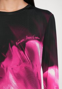 Majica z dolgimi rokavi, ki združuje črno tkanino z živahnim roza abstraktnim vzorcem in logotipom "Versace Jeans Couture" v roza pisavi.