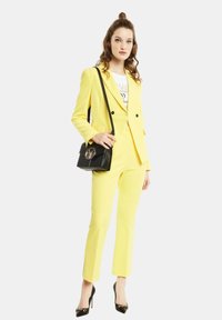 Completo giallo su misura con giacca strutturata e pantaloni slim. Borsa nera con dettaglio del logo e tacchi a punta neri. T-shirt bianca con grafica sotto.