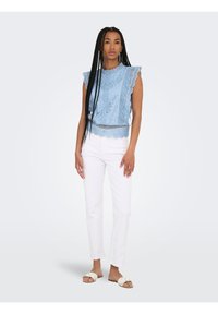 Femme aux longues tresses portant un haut sans manches en dentelle bleu clair, un pantalon blanc et des sandales beiges à enfiler, debout devant un fond clair.