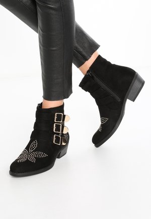 Schwarze Wildleder-Stiefeletten mit drei goldenen Schnallen, seitlichem Reißverschluss und genietetem Blumenmuster, getragen zu schwarzen Lederhosen.