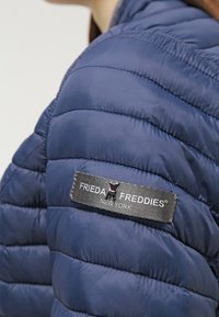 Giacca puffer trapuntata blu navy con design cucito, texture liscia e etichetta in tessuto con "FRIEDA FREDDIES® NEW YORK" in grigio.