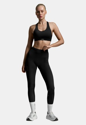 Kobieta stojąca, ubrana w czarny sportowy biustonosz i legginsy, białe skarpetki oraz białe tenisówki na jednolitym tle.