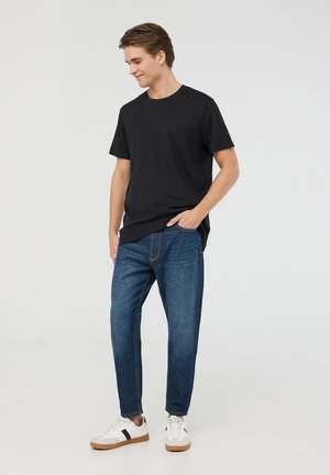 T-shirt in cotone nero, vestibilità rilassata, abbinato a jeans di denim azzurro chiaro. Sneakers bianche con accento a strisce nere. Outfit semplice e casual.