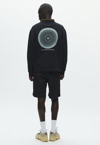 Sweat-shirt noir avec un graphisme circulaire au dos, accompagné d'un short noir. Le mannequin porte des chaussettes blanches et des baskets beiges.