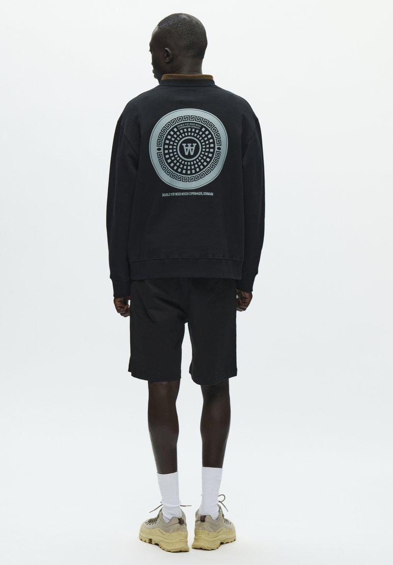 Sweat-shirt noir avec un graphisme circulaire au dos, accompagné d'un short noir. Le mannequin porte des chaussettes blanches et des baskets beiges.