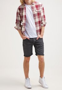 Homme portant une chemise en flanelle rouge à carreaux par-dessus un t-shirt blanc, un short foncé retroussé, et des baskets montantes blanches, debout avec les mains dans les poches.