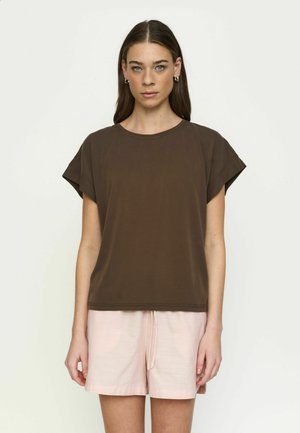 Kvinde med langt brunt hår iført en brun kortærmet skjorte og lyserøde shorts, stående mod en ensfarvet lys baggrund.