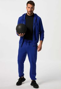 Sudadera deportiva azul y pantalones de chándal de tela suave, combinados con una camiseta negra y zapatos deportivos negros, sosteniendo un balón de baloncesto negro.