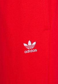 Červená látka ze směsi bavlny s malým bílým logem Adidas, které má design tří listů a horizontální pruhy. Hladká textura, přiléhavý střih.