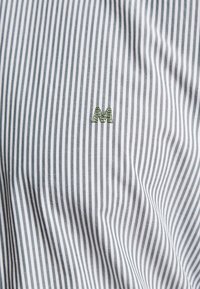 Tessuto a righe grigie e bianche con una texture morbida, caratterizzato da un piccolo logo "M" ricamato di colore verde.