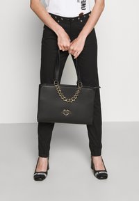 Svart handväska med en strukturerad design, guldhjärta-formad kedjeaccent och "LOVE MOSCHINO"-logotyp på framsidan. Slät, texturerad yta.