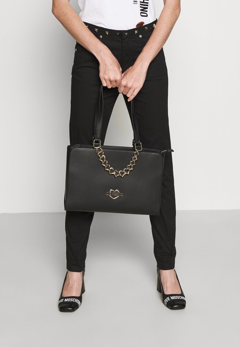 Svart handväska med en strukturerad design, guldhjärta-formad kedjeaccent och "LOVE MOSCHINO"-logotyp på framsidan. Slät, texturerad yta.
