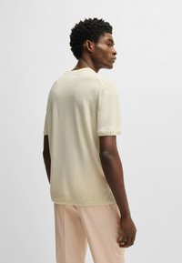 Helles beigefarbenes Strick-T-Shirt mit kurzen Ärmeln, Rundhalsausschnitt und geripptem Saum. Es hat eine weiche Textur und einen leicht entspannten Schnitt und wird mit hellen Hosen getragen.