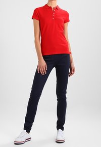 Röd pikétröja med krage och korta ärmar, parat med marinblå slim-fit byxor och vita sneakers med färgade ränder.