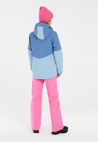Giacca da sci impermeabile in blu scuro e chiaro, abbinata a pantaloni da sci rosa brillante. Berretto a maglia rosa, stivali da neve neri resistenti.