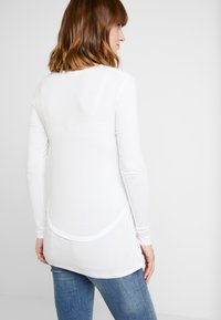 Camisa de manga longa branca feita de tecido macio, com uma bainha arredondada, ajuste slim e design minimalista. Usada sobre jeans azuis casuais.