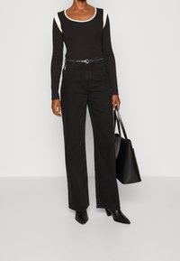 Schwarzes Langarmoberteil mit cremefarbenen Akzenten, hochgewaist schwarze Jeans, schwarzer Gürtel und schwarze, spitze Absatzstiefel. Große schwarze Tote-Tasche.
