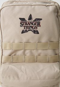 Bežová látková taška s logom "Stranger Things" v čiernej a červenej farbe, s horizontálnymi popruhmi pre ďalšie upevnenie.