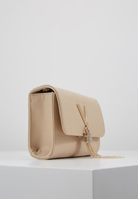Beige handtas met textuur en klep sluiting, met een accent van goudkleurig beslag en een kettingriem. Compact rechthoekige vorm.