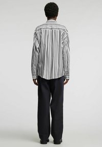 Koszula w paski w czerni i bieli, w oversize’owym kroju, z kołnierzykiem zapinanym na guziki i długimi rękawami. Noszona z szerokimi granatowymi spodniami i czarnymi butami.