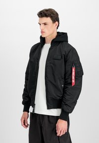 Black bomber jacket with a hood, zip closure, ribbed cuffs, and a red tag on the left sleeve; made of a smooth, shiny material.