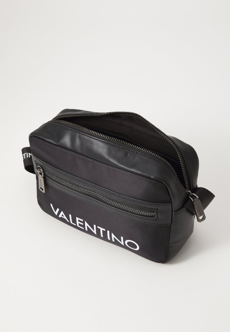 Sac bandoulière en tissu noir avec un dessus en cuir lisse, des fermetures éclair et un logo "VALENTINO" blanc ; comprend une poche latérale avec fermeture éclair.
