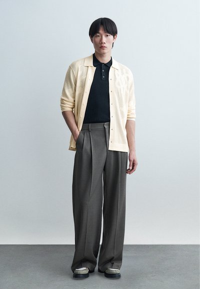 Νέος άνδρας στέκεται με τα χέρια στις τσέπες, φοράει ανοιχτό μπεζ ζακέτα, μαύρο πόλο μπλουζάκι, φαρδιά γκρι παντελόνια και σκούρα παπούτσια.