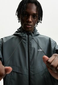Siva zip-up jopič z kapuco, ki ima vrvico in bel logotip na prsih. Gladka tekstura s lahko, športno zasnovo.