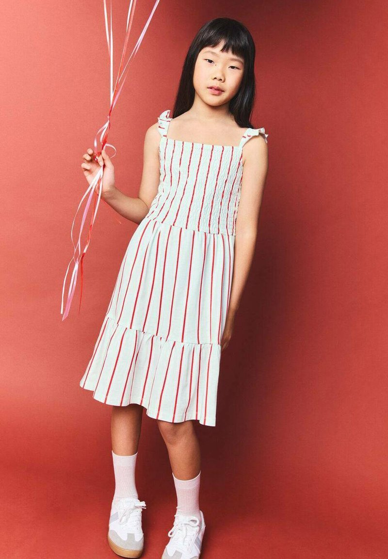 Robe blanche à rayures rouges verticales, encolure carrée et bretelles à volants. Comporte une jupe à volants. Associée à des baskets blanches et des chaussettes.