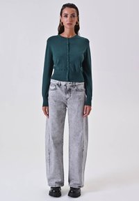 Grüne Strickjacke mit Rundhalsausschnitt und Knopfverschluss, kombiniert mit hochgeschnittenen, hellgrauen weiten Jeans und schwarzen Plateaustiefeln.