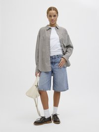 Camisa às riscas com botão, t-shirt branca por baixo, calções de ganga azuis, meias brancas, sapatos castanhos com atacadores, segurando uma mala texturizada de cor creme.