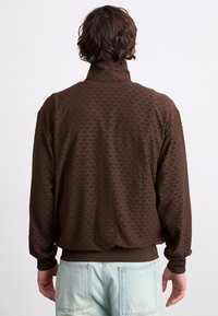 Καφέ φleece ζακέτα με φερμουάρ που διαθέτει υφή φύλλων, ψηλό γιακά και ριμπ μανσέτες. Φοριέται με ανοιχτόχρωμα μπλε τζιν παντελόνια.