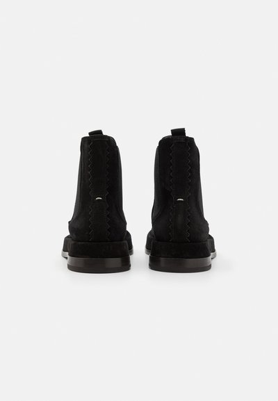 Botine negre din suede, cu textură netedă, panouri laterale elastice și accente ondulate. Prezentând un toc jos și îngroșat și un tab de prindere întărit pe spate.