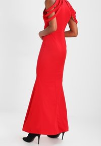 Abito da sera rosso con silhouette aderente, design a spalle scoperte e dettagli a cinturino, orlo svasato. Indossato con tacchi neri.