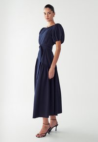 Marineblauwe midi-jurk met pofmouwen, een getailleerde taille en een gelaagde rok. Glad materiaal, minimale details, gecombineerd met zwarte bandjeshakken.