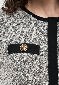 Close-up of black and white textured knit garment with black trim and gold button detail on chest pocket area.