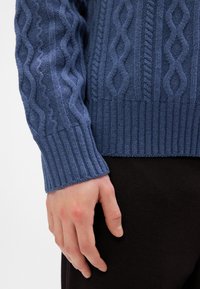 Blauwe gebreide trui met een getextureerd kabelpatroon en geribbelde manchetten en zoom. Een hand steunt tegen een donkere stof eronder.