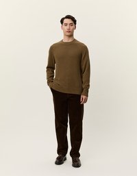 Καφέ πλεκτό πουλόβερ με ριγέ μανίκια, συνδυασμένο με σκούρες καφέ βελουτέ παντελόνες. Το μοντέλο στέκεται ακριβώς μπροστά σε ουδέτερο φόντο.