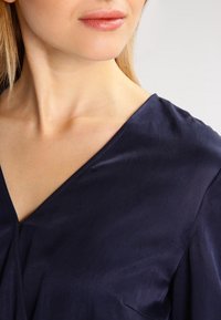 Námorně modrá blůza s výstřihem do V, vyrobená z jemné tkaniny, s měkkým drapováním a jemným leskem, zdůrazňující uvolněný střih.