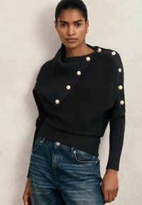 Femeie îmbrăcată cu un pulover negru texturat, cu butoane aurii pe umăr și pe brațe, purtând blugi albaștri, stând în fața unui fundal simplu.